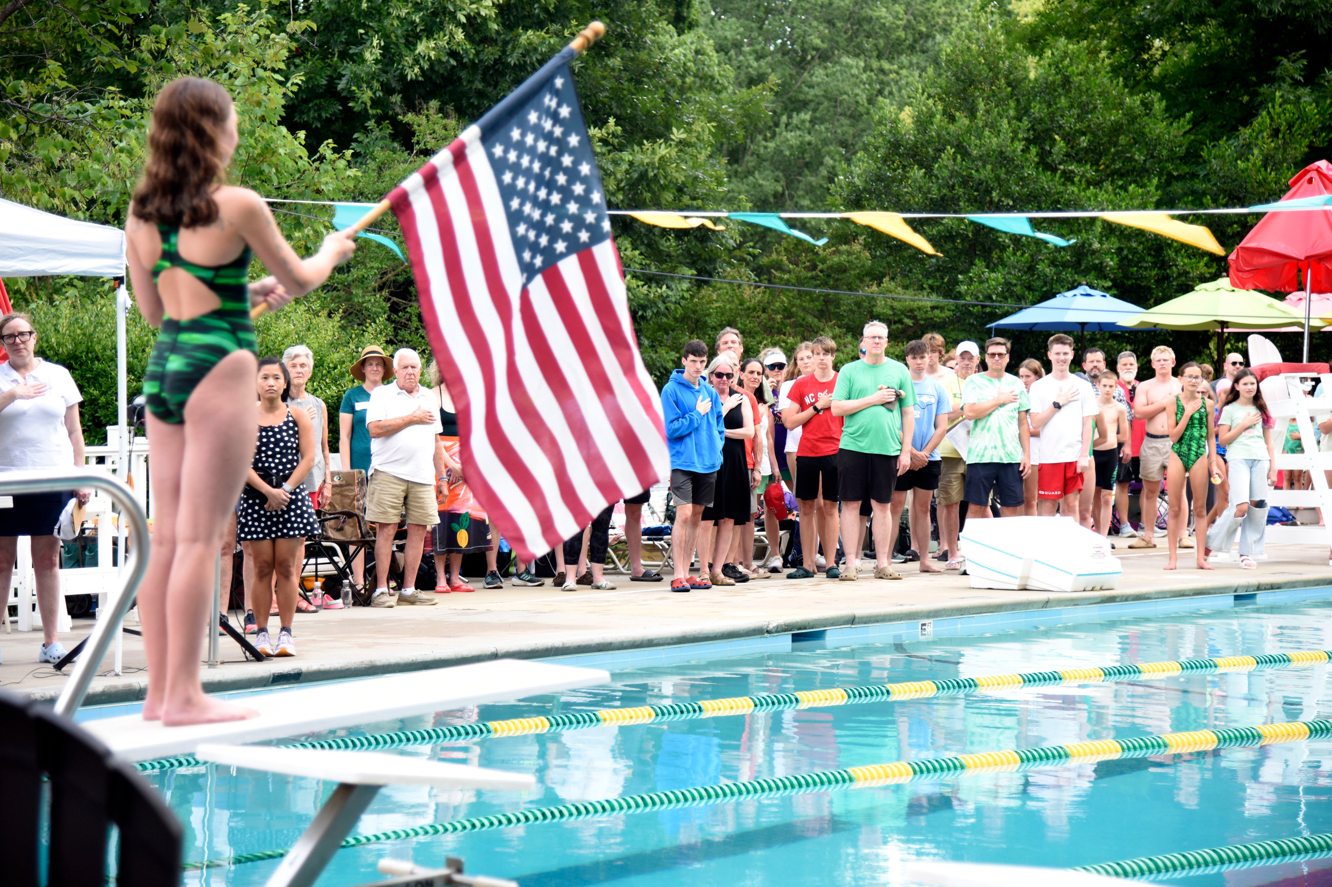 A photo of Friendly swimmer Frankie Hogan holding the American flag on the pool diving board during the National Anthem at a home swim meet. Families and swimmers are on the deck with hands over their hearts.
